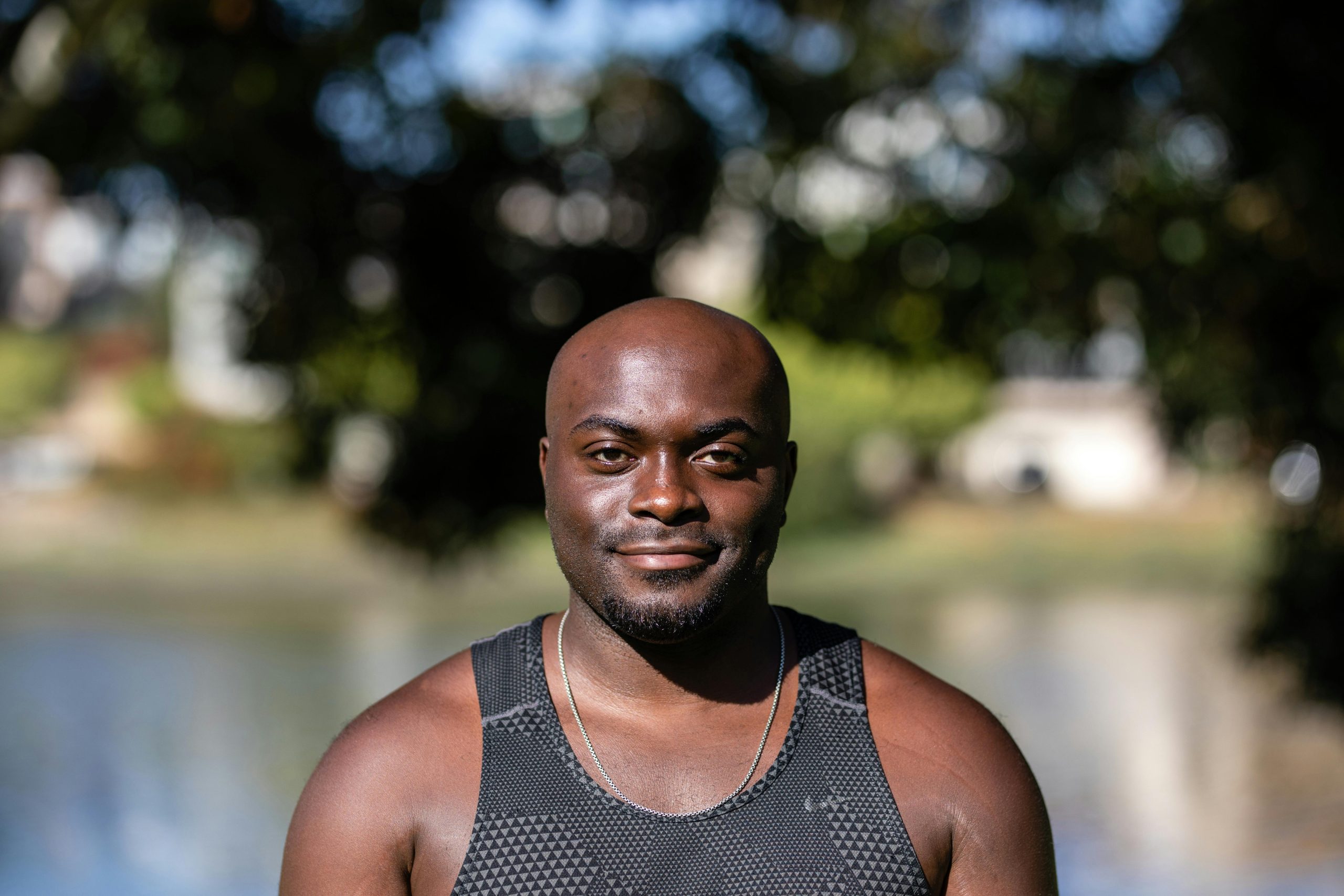Portrait of a smiling bald African American man in a black tank top outdoors with a blurred background.
